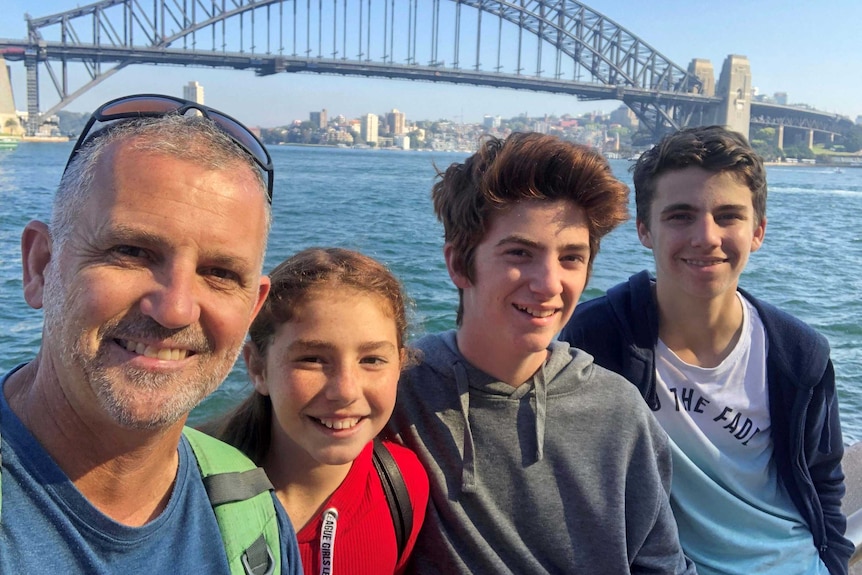 A dad and his three teenaged kids are smiling in front of the Sydney Harbour Bridge on a sunny day.