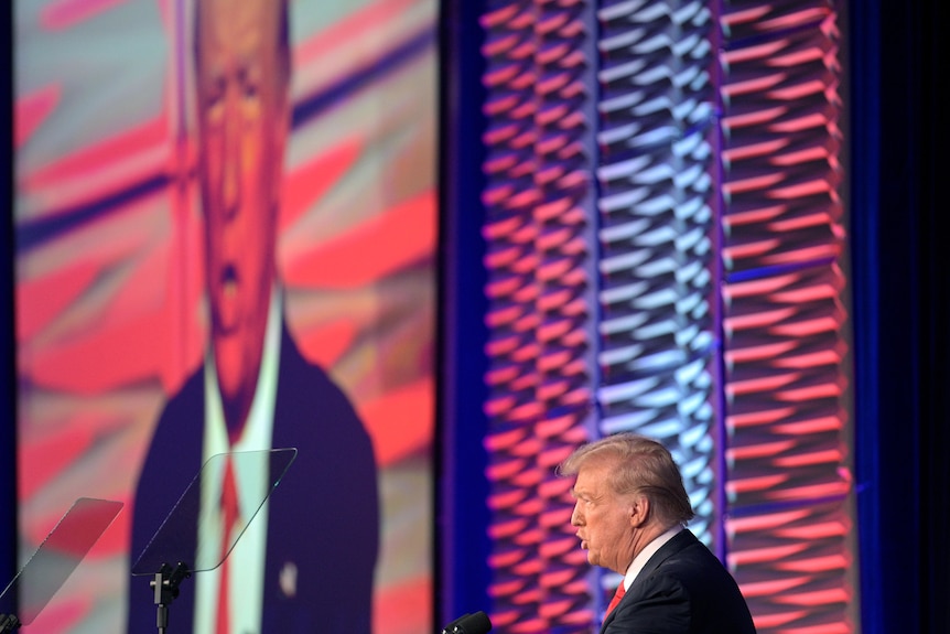 Donald Trump speaks on stage with a huge TV image of him behind him