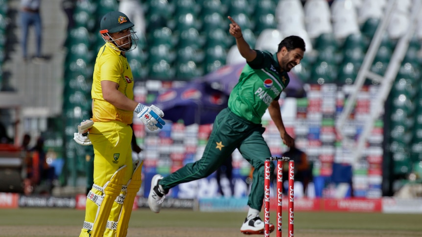 An Australian ODI cricketer looks dejected after being given out as a bowler runs away with arms out in celebration.