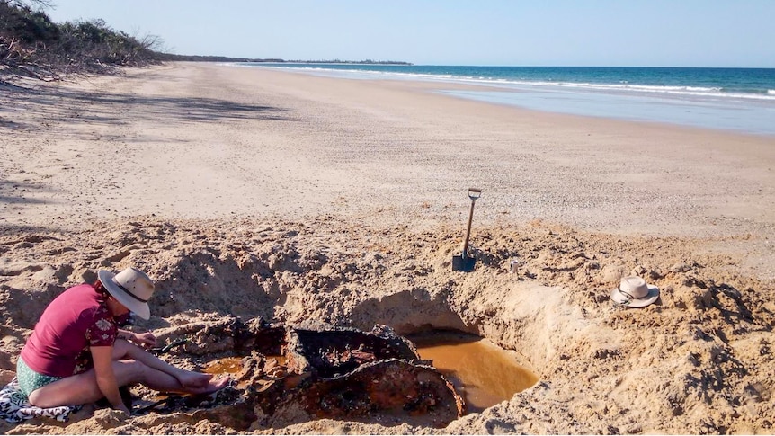 A woman digs around a rusty car body on a beach.