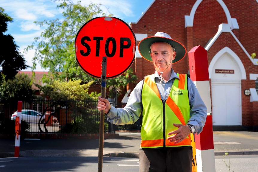 School crossing guard Robert Stove stands in the middle of the road with a crossing sign in front of a school.