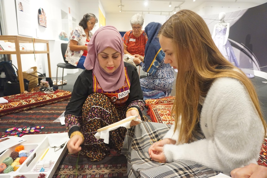 An Afghan woman wearing a pink headscarf demonstrates embroidery to a younger white woman. They sit on the floor.