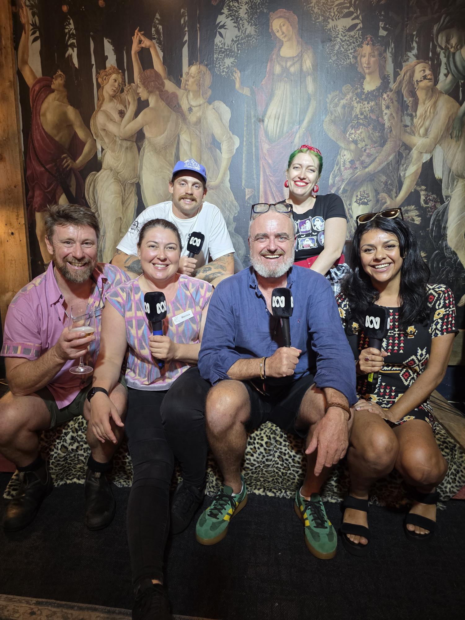 Six smiling people hold ABC microphones in front of a classical painted mural.
