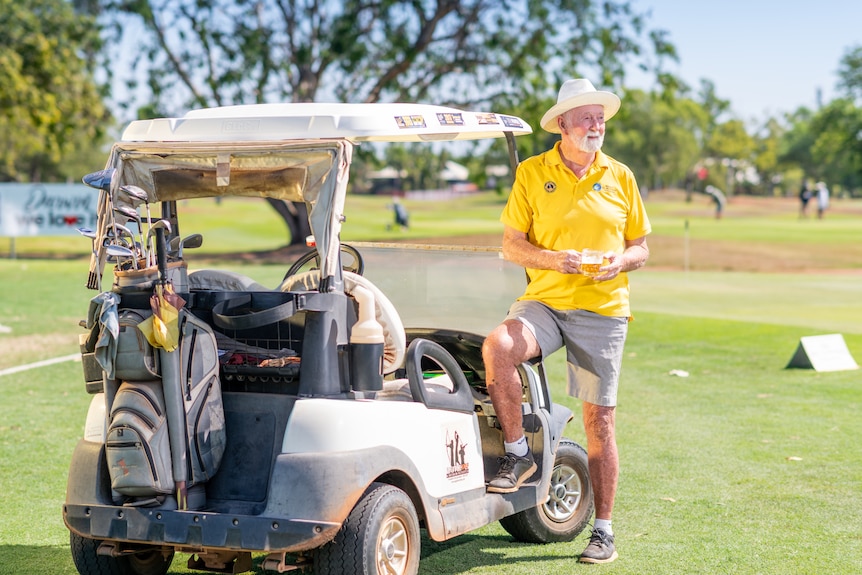 Elderly man stands next to a golf buggy smiling on a sunny day.