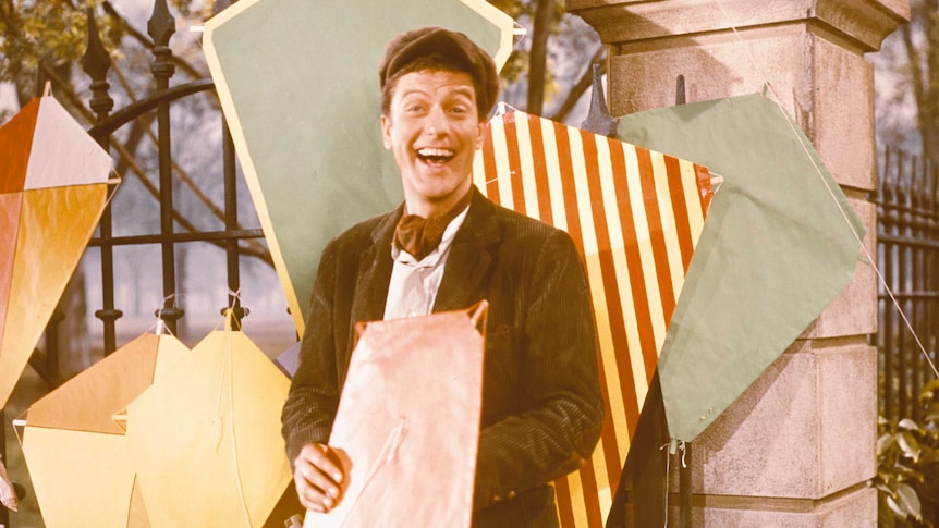 Dick Van Dyke holds a kite and smiles at the camera on the set of Mary Poppins. Kites adorn a fence behind him.