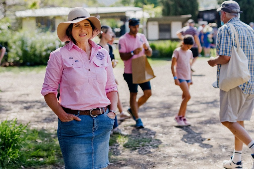 A smiling woman in a denim skirt, pink shirt, an akubra stands with hands in pocket as people mill around in an open grounds.