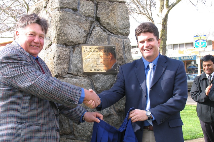 Two men shaking hands in front of a golden plaque attached to a rocky sculpture.