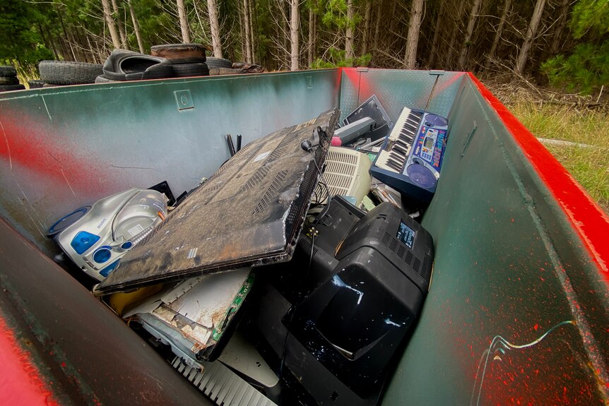 A skip filled with electrical waste including dirty televisions, a keyboard, and computers can be seen. 