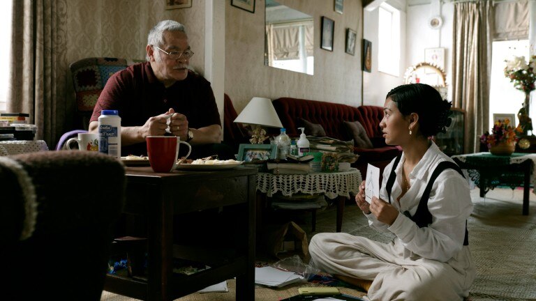 Actress Luciane sits on the floor during a scene with actor Albert Rounds who is sitting on a couch. 