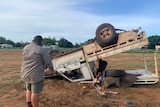 A ute tipped over and laying on its roof is in wreckage, next to it Dan Hanley looks at the wreck.