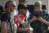 Three men wearing sunglasses and camouflage vests and one holding a gun watch a crowd.