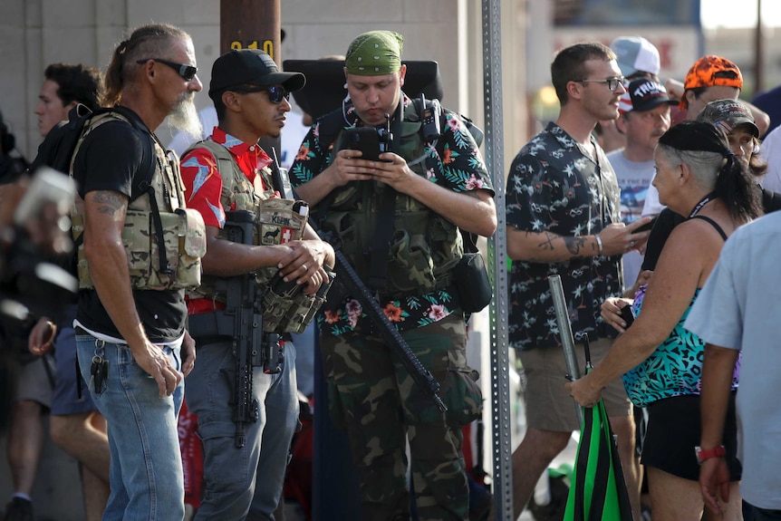 Three men wearing sunglasses and camouflage vests and one holding a gun watch a crowd.