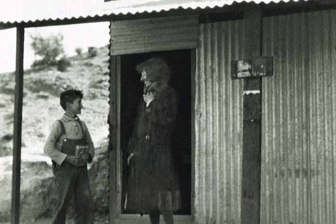 A tall woman wearing a long warm coat stands under the corrugated iron verandah of a store talking to a child.