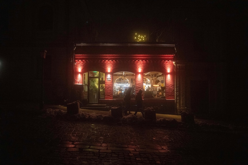 People walk past a cafe with red and yellow lights shining against a dark night.
