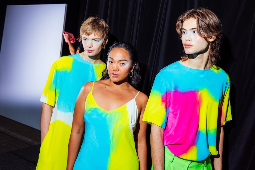 A non-binary white person, a woman of colour and a queer white person model vibrant hand-painted clothing against a dark curtain