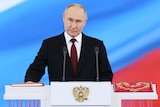 Vladimir Putin holds his hand on a red book in front of a screen showing the Russian flag colours of red, white and blue