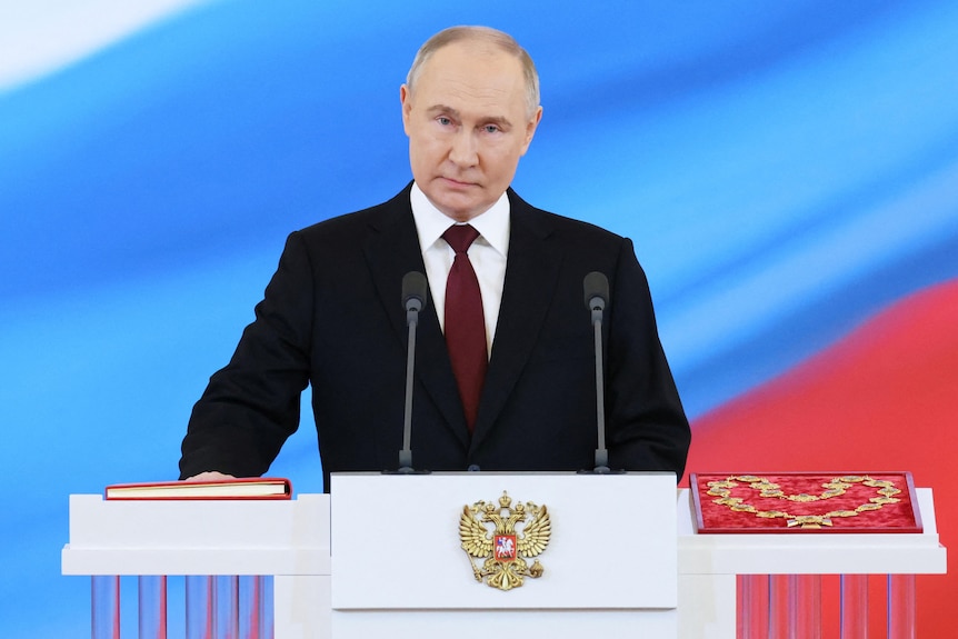 Vladimir Putin holds his hand on a red book in front of a screen showing the Russian flag colours of red, white and blue