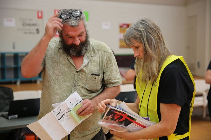 Bearded man with greying hair and reading glasses on his head holds papers and looks at papers being shown to him by a woman