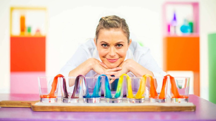 Rachael on the Play School Science Time set wearing a lab coat standing behind drinking glasses with coloured paper