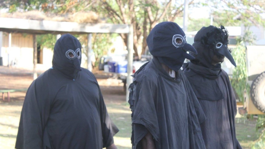 Three people in black, beaked bird costumes walk across a school oval