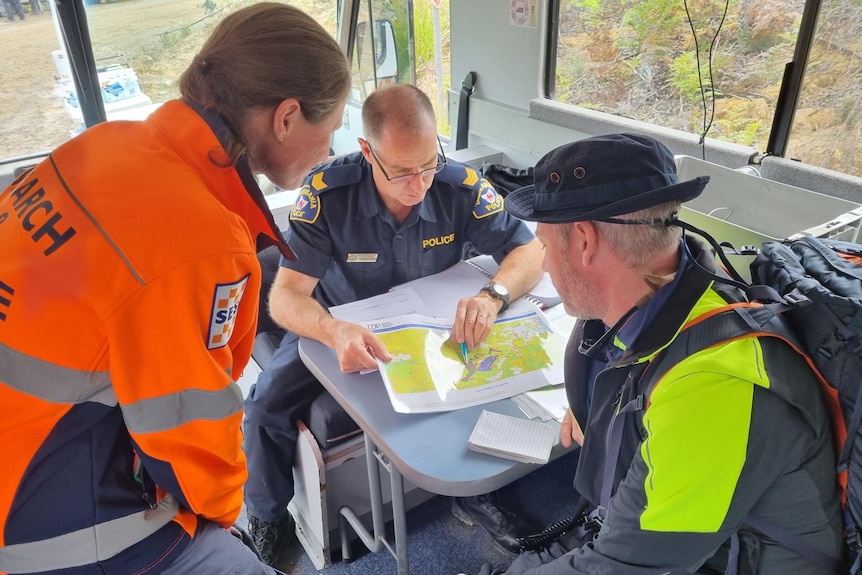 Two search and rescue workers in hi-vis leans over a map. A seated Tasmania Police officer points at the map. 