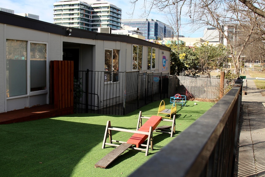 Empty play equipment sits outside a childcare centre on fake grass behind a fence