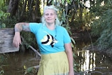 a woman in blue t shirt with a fish on it, leaning on one hand on a tree branch in front of small creek