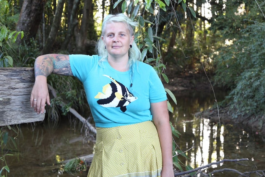 a woman in blue t shirt with a fish on it, leaning on one hand on a tree branch in front of small creek