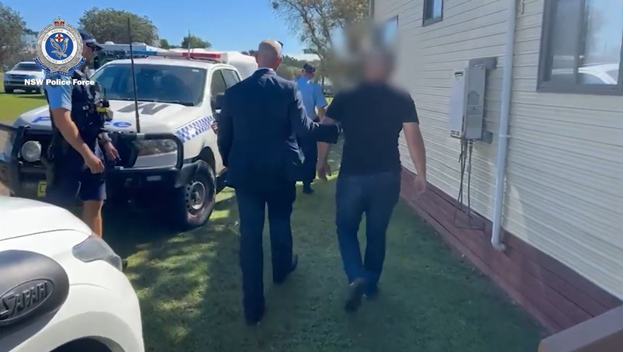Uniformed and planinclothes police escort a man towards a police vehicle in a caravan park.