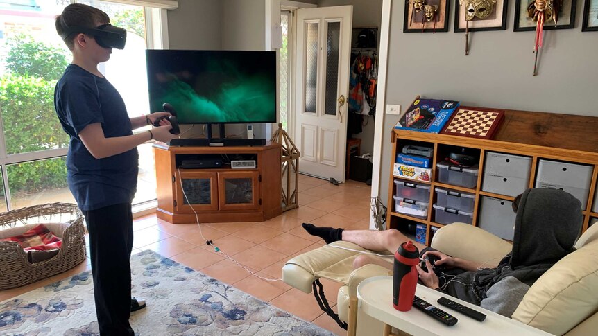 Two teenage boys in a living room playing video games. One is standing, wearing a VR headset
