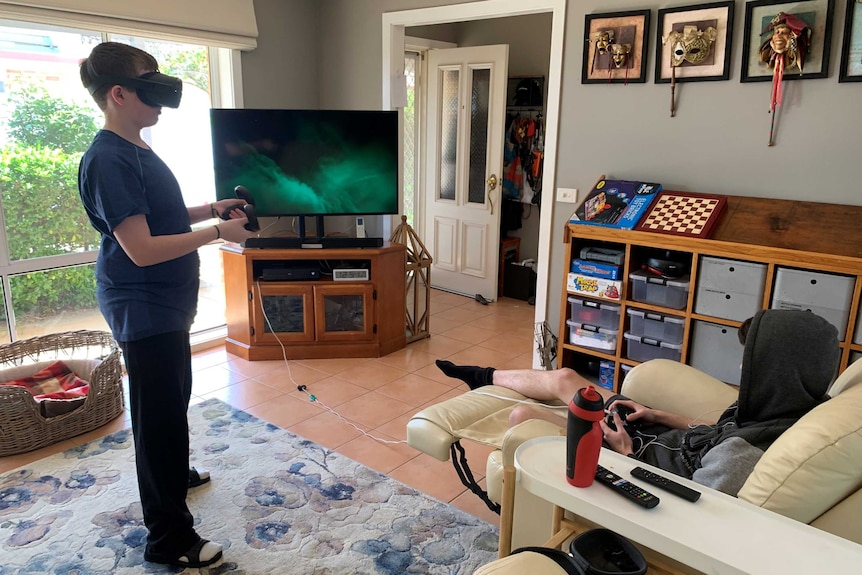 Two teenage boys in a living room playing video games. One is standing, wearing a VR headset