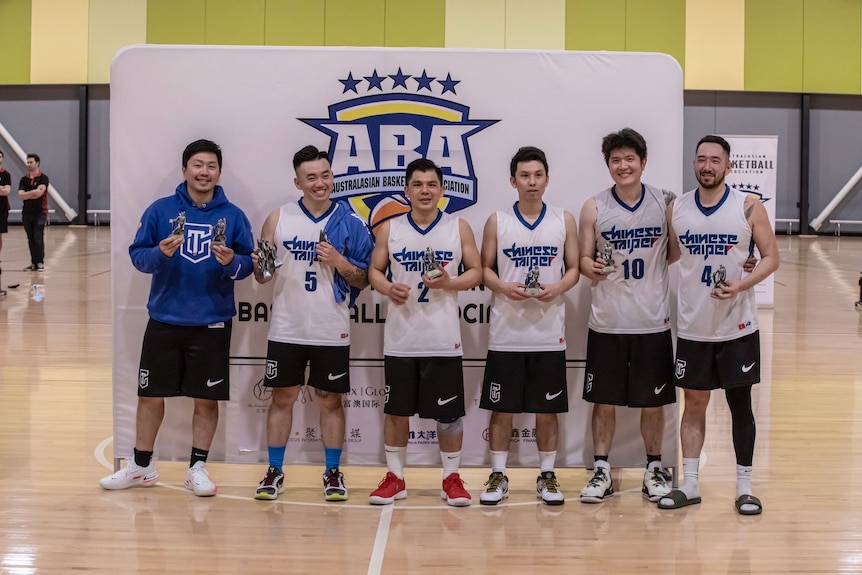 Six men in basketball gear standing in front of a banner on a court.