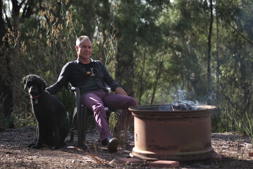The writer Kim Scott in his backyard with his dog and a fire pit, bush in the background