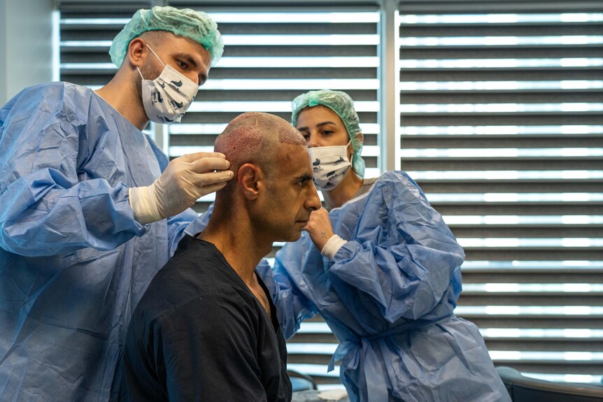 A man wearing blue hospital gown sits up while nurses in scrubs inspect his scalp