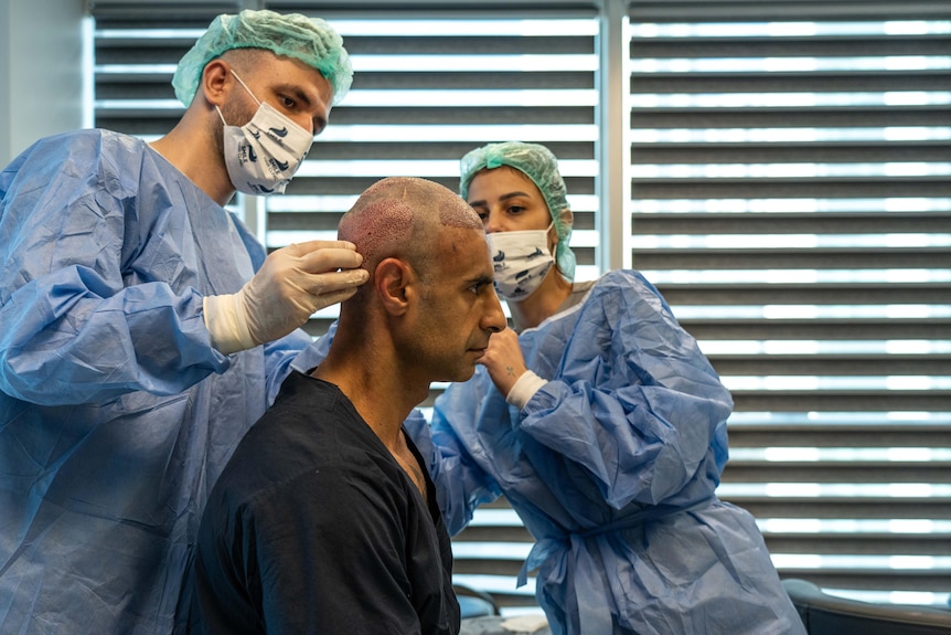 A man wearing blue hospital gown sits up while nurses in scrubs inspect his scalp