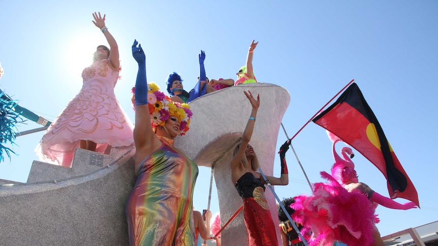 Seven very colourful and shiny drag queens wave from atop a giant silver high heel. One waves an Indigenous flag.