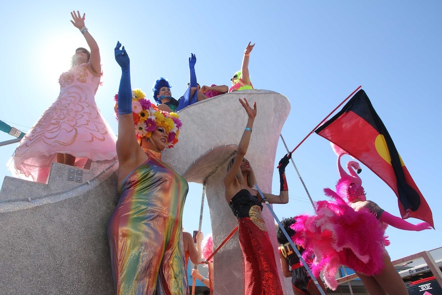 Seven very colourful and shiny drag queens wave from atop a giant silver high heel. One waves an Indigenous flag.