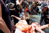 A woman with brown curly hair and in a green jacket is being dragged from the waters by a police officer while she holds a si