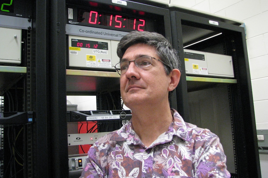 A man with glasses standing in front of atomic clock, which has digital time face.