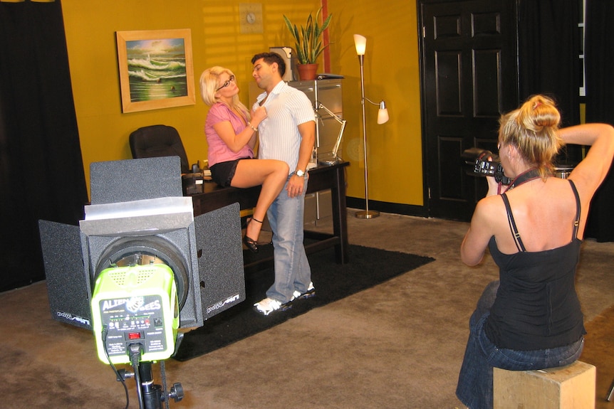 A man and woman are on set of a pornographic film and another woman is standing behind a video camera