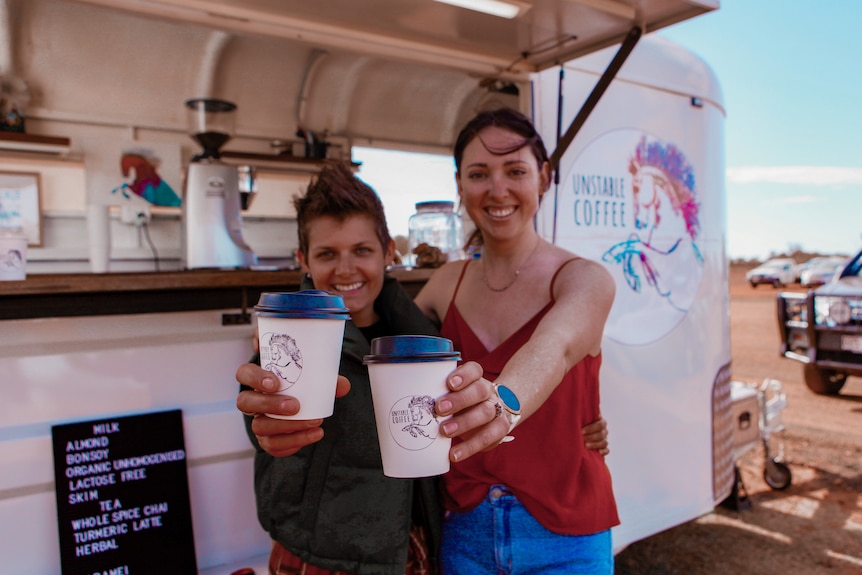 Two woman hold coffee cups in front of a coffee van made from a horse float