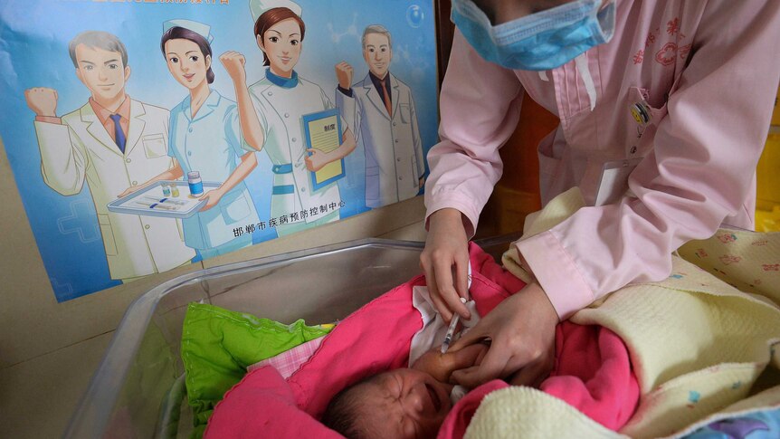 A nurse wearing a face mask leans over a cot to give a baby a vaccine shot.