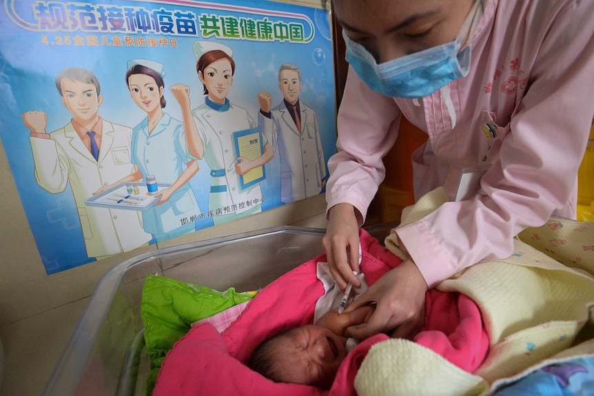 A nurse wearing a face mask leans over a cot to give a baby a vaccine shot.