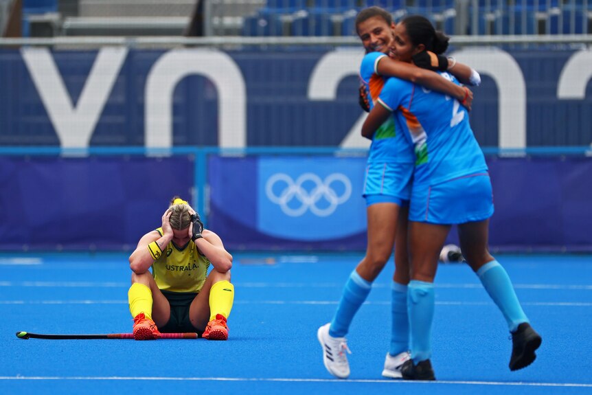 A group of Indian hockey players in blue celebrate as a single yellow player sits and looks sad in a hockey match.