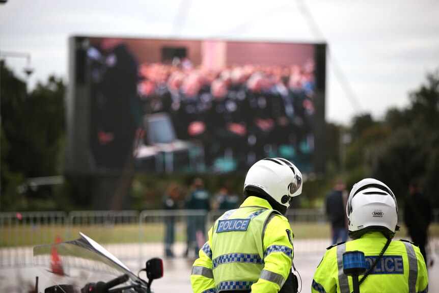 Two WA Police officers in hi vis vests and white motorbike helmets stand in front of a big screen outside Perth Stadium.