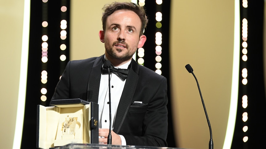 Man in black tux on stage holding award, looking upwards.