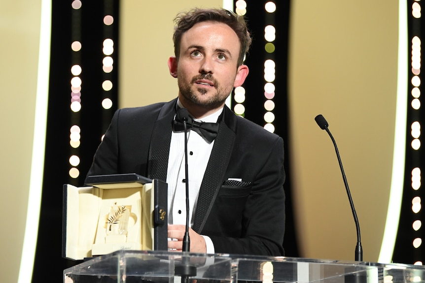 Man in black tux on stage holding award, looking upwards.