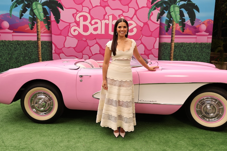 America Ferrera in a white dress standing in front of a pink Barbie convertible with Barbie written behind her