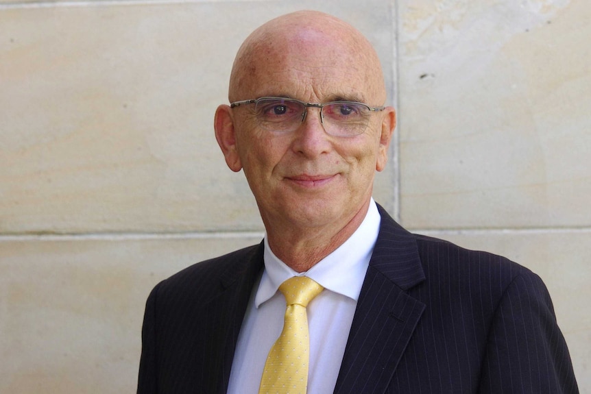 A bald, bespectacled man in a dark suit with a gold tie.