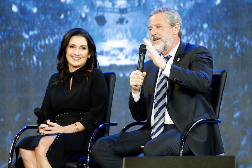 A man seated on stage with a microphone gestures alongside a woman who smiles, also holding a microphone.